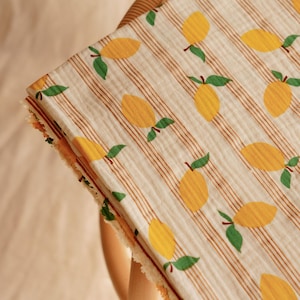 May include: A folded, cream-colored fabric with a repeating pattern of yellow lemons and green leaves. The fabric has thin, vertical brown stripes and a frayed edge. The fabric is draped over a wicker chair.