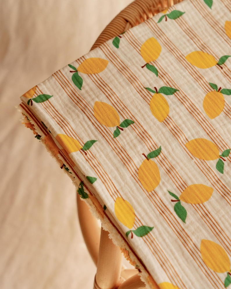 May include: A folded, cream-colored fabric with a repeating pattern of yellow lemons and green leaves. The fabric has thin, vertical brown stripes and a frayed edge. The fabric is draped over a wicker chair.