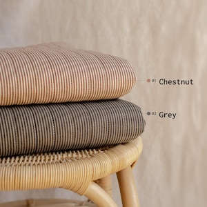 May include: Two folded fabric cushions with a vertical pinstripe pattern. The top cushion is chestnut brown with white stripes, labeled 01 Chestnut. The bottom cushion is grey with white stripes, labeled 02 Grey. Both rest on a light-colored rattan stool.