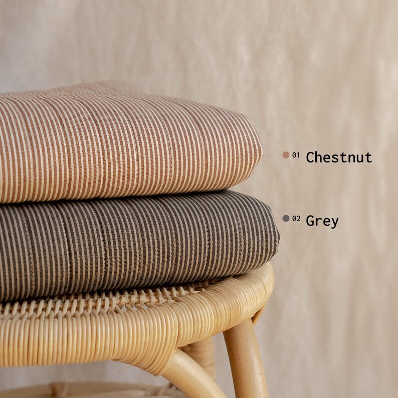 May include: Two folded fabric cushions with a vertical pinstripe pattern. The top cushion is chestnut brown with white stripes, labeled 01 Chestnut. The bottom cushion is grey with white stripes, labeled 02 Grey. Both rest on a light-colored rattan stool.