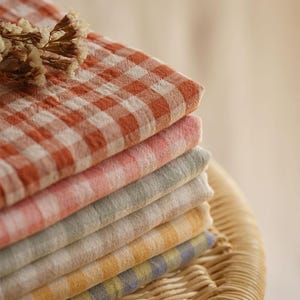 May include: A stack of colorful, checkered fabric cloths in shades of orange, pink, green, yellow, and blue. The cloths are neatly folded and stacked on top of each other. A sprig of dried flowers rests on top of the stack. The cloths are on a wicker surface.