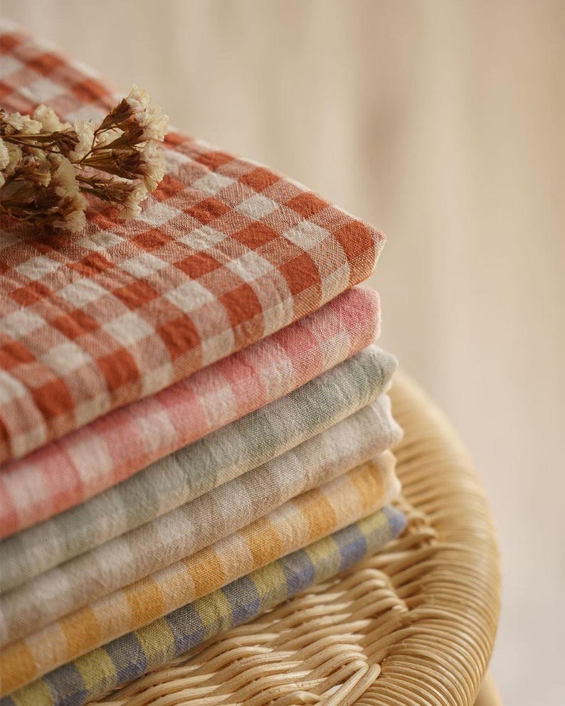 May include: A stack of colorful, checkered fabric cloths in shades of orange, pink, green, yellow, and blue. The cloths are neatly folded and stacked on top of each other. A sprig of dried flowers rests on top of the stack. The cloths are on a wicker surface.