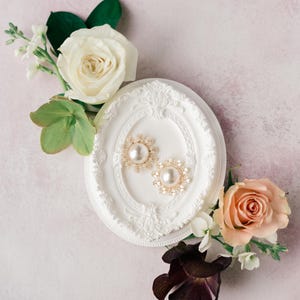 以下が含まれることがあります： 金色のイヤリング、中央にパールと花の装飾が施されています。白い装飾的な楕円形のフレームに展示されています。イヤリングは白と桃色のバラ、緑の葉に囲まれ、ピンク色の背景に配置されています。