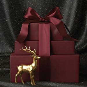 Puede incluir: Una pila de cajas de regalo de color burdeos atadas con una gran cinta y lazo burdeos. Una figura de ciervo dorada se encuentra frente a los regalos. El fondo es una tela oscura y brillante.