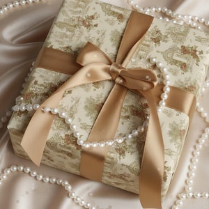 Op de afbeelding: Een cadeau verpakt met een crèmekleurig bloemenpatroon, vastgebonden met een gouden lint en strik. Een streng witte parels is om het cadeau gedrapeerd, wat een elegante presentatie creëert. Het cadeau ligt op een satijnen oppervlak.