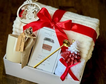May include: A white gift box filled with a white towel, a mug, a bag of hot chocolate mix, a small wooden snowman ornament, a gold spoon, and a vanilla latte mix packet. The box is tied with a red ribbon and bow.