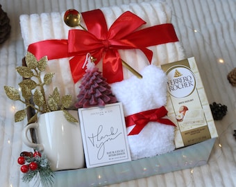 May include: A white gift basket filled with a white throw blanket, a white mug, a white fluffy slipper, a red candle shaped like a Christmas tree, a box of white chocolate Ferrero Rocher, and a small card that says "Thank You". The basket is decorated with a red ribbon and a gold sprig of leaves.