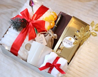May include: A gift basket filled with a white blanket, a red bow, a white mug, a pen, a notebook, a box of chocolates, a small towel with a red bow, and a small bag with a tag that says "TEA".