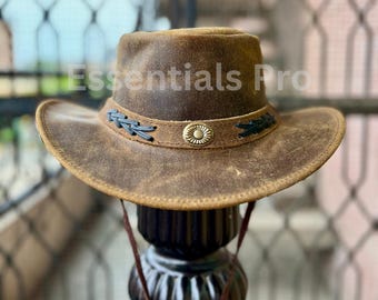 Puede incluir: Un sombrero vaquero de cuero marrón con una banda trenzada negra y un acento de metal dorado. El sombrero tiene un aspecto desgastado y está usado.