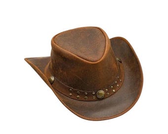May include: A brown distressed leather cowboy hat featuring a pinched crown and a wide, curved brim. The hat is accented with a matching leather band adorned with small metallic studs and a central circular metal concho detail on the front.