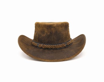 Puede incluir: Un sombrero de vaquero de cuero marrón con acabado desgastado, que presenta un ala ancha y una copa con una ligera hendidura. El sombrero está decorado con una banda compuesta por una tira de cuero trenzado y tachuelas de metal oscuro.