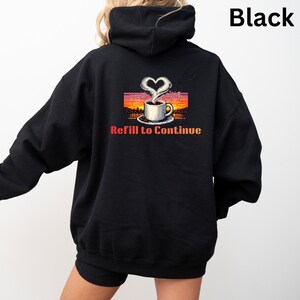 Peut inclure: Sweat à capuche noir avec un motif de tasse à café. Le motif représente une tasse de café avec de la vapeur en forme de cœur, sur fond de coucher de soleil. Le texte "Refill to Continue" est en dessous.
