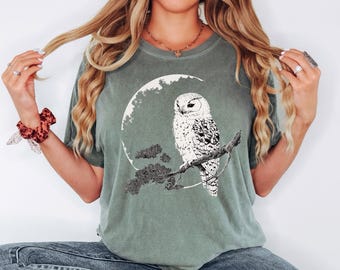 May include: A green t-shirt with a white owl perched on a branch in front of a crescent moon. The owl has black eyes and a detailed pattern of feathers.