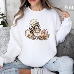 Puede incluir: Una sudadera blanca de manga larga con una ilustración gráfica de un mapache con gorro de chef y delantal, mezclando ingredientes en un bol rodeado de harina, azúcar, huevos y galletas. La palabra WHITE está impresa en la esquina superior.