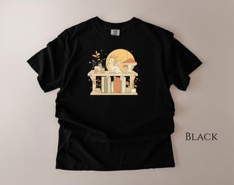 May include: Black t-shirt featuring a whimsical illustration. The design includes a white dragon, mushrooms, and books on a shelf, set against a yellow moon. The shirt is a comfortable, casual wear.