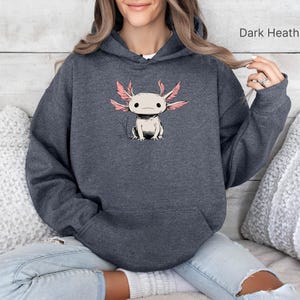 May include: Dark heather grey hoodie with a cartoon axolotl design. The axolotl has pink gills and is white and grey. The hoodie has a front pocket and a drawstring hood. The text "Dark Heather" is visible.