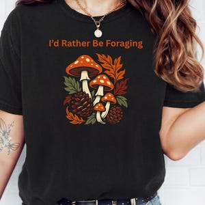 May include: Black t-shirt with the text "I'd Rather Be Foraging" in orange. The shirt features a colorful illustration of mushrooms, leaves, and pinecones in shades of orange, brown, and green.