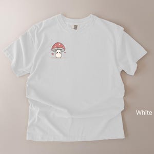 May include: White t-shirt featuring a small, cute mushroom graphic with a red and white cap and a smiling face. The mushroom is surrounded by small hearts. The word "White" is printed on the bottom right corner of the image.