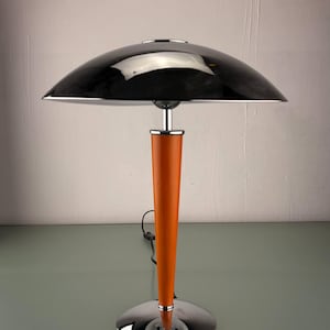 May include: A modern table lamp with a black, dome-shaped shade and a chrome base. The lamp features a vibrant orange stem. The design is sleek and contemporary, suitable for various interior styles. The lamp's height is approximately 50 cm.
