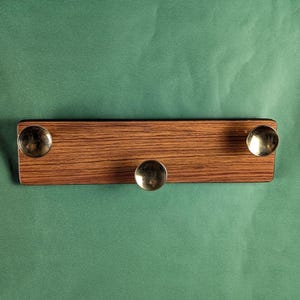 Op de afbeelding: Een houten wandbevestigde sleutelhanger met drie messinghaken. Het hout heeft een donkerbruine vlek en een houtnerfpatroon.
