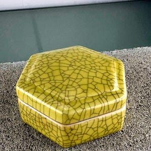 May include: A hexagonal, lime green ceramic box with a cracked glaze pattern. The box has a lid and a white rim. The box is sitting on a gray surface.