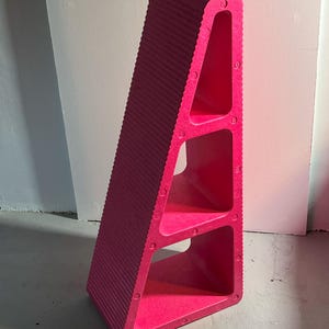 May include: A vibrant pink, angled shelving unit with three open compartments. The exterior has a ribbed texture, and the unit is made of a solid material. The shelf is leaning against a white wall.