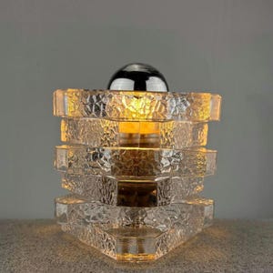 May include: A clear glass table lamp with a textured, stacked design. The lamp has a chrome dome on top and emits a warm, yellow light. The lamp's dimensions are approximately 8 inches tall and 6 inches wide.
