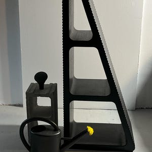 以下が含まれることがあります： 注ぎ口に黄色い花がついた黒いジョウロと、3段の棚が付いた黒い三角形の棚ユニットと、灰色のコンクリートブロック