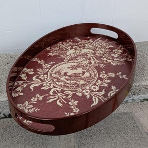 Peut inclure: Un plateau de service ovale avec une base rouge foncé et un motif floral blanc. Le plateau a deux poignées sur les côtés.