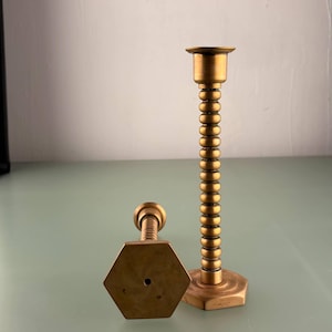 May include: Two brass candle holders. One is upright with a beaded stem and a hexagonal base. The other is lying down, also with a hexagonal base and a screw-like top. The candle holders are on a light green surface.