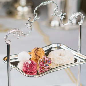 Puede incluir: Un soporte de servicio de plata de dos niveles con un diseño decorativo de desplazamiento. El soporte está lleno de una variedad de galletas y dulces coloridos y blancos.