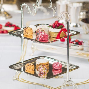 May include: A silver two-tiered serving stand with ornate details. The top tier holds a variety of small cakes and pastries, including a cake with red berries and a red ribbon.