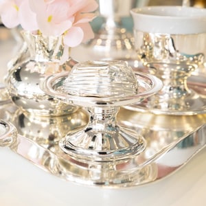 Könnte beinhalten: Ein silbernes Serviertablett mit einem kleinen silbernen Fußschälchen mit strukturierter Abdeckung. Eine silberne Vase mit rosa Blumen und eine silberne Teetasse befinden sich ebenfalls auf dem Tablett. Die Artikel sind reflektierend und elegant.