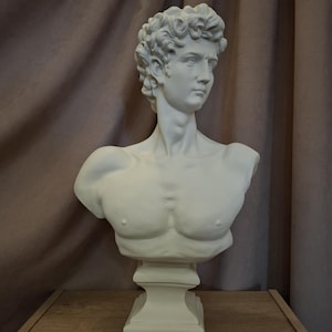 May include: White plaster bust sculpture of a man with short, curly hair. The bust is on a white pedestal.