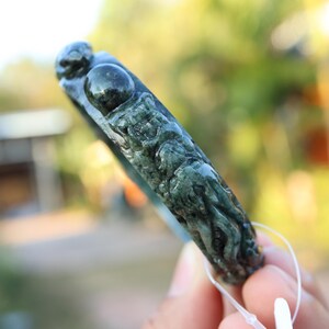 May include: Dark green jade bracelet with carved relief details. The bracelet features a detailed carving of a mythical creature. The jade has a polished finish and is held by a white string.