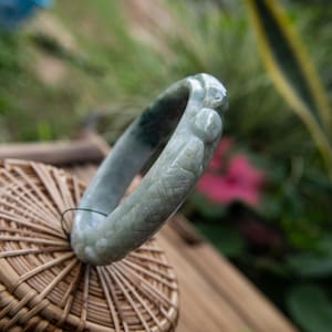 May include: A pale green jade bangle bracelet with carved detailing. The bracelet has a smooth, polished finish and a slight colour variation. The design includes carved patterns and a rounded shape, suggesting a traditional jewellery style.