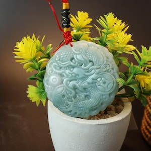 May include: A round jade pendant featuring an intricate carving of a dragon and fish. The pendant is pale green and has a red and gold cord attached.