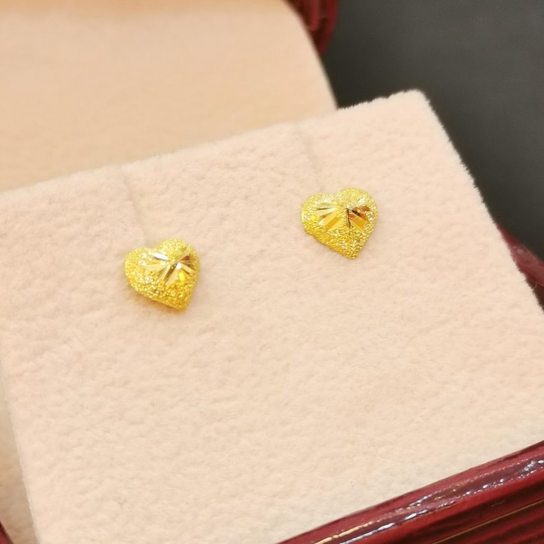 May include: A pair of gold heart-shaped earrings with a textured surface. The earrings are displayed in a velvet-lined box.