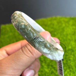 May include: A jade bangle bracelet with carved floral designs. The bracelet features a mix of white and green hues, with the carved details in a darker green. The bracelet is held against a green background.