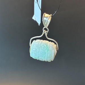 May include: A jade pendant with intricate carvings, suspended from a silver bail. The pendant is shaped like a cylinder and has a light green colour.