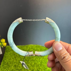 May include: A light green jade bangle bracelet with silver accents. The bracelet is held together by a silver chain and clasp. The jade has a marbled appearance. The bracelet is being held up by a hand.