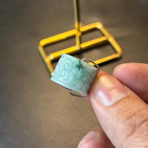 May include: A green jade ring with a carved design. The ring is set in silver and is on a finger.