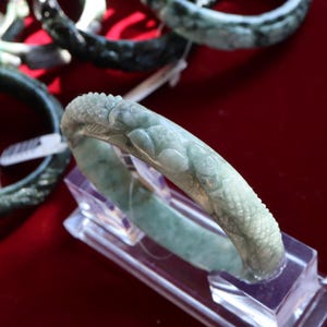May include: A pale green jade bangle bracelet featuring carved dragon and scale patterns. The bracelet is displayed on a clear stand, with other jade bangles visible in the background. The jade has a translucent quality.