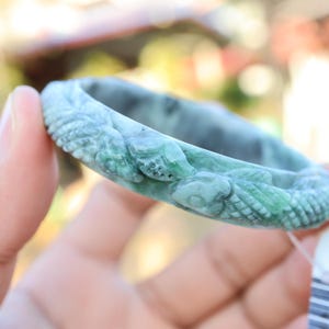 May include: A jade bangle bracelet with a carved design. The bracelet is a mix of green and grey hues, with detailed carvings of what appear to be mythical creatures. The bracelet is held in a hand, showcasing its intricate details and polished finish.