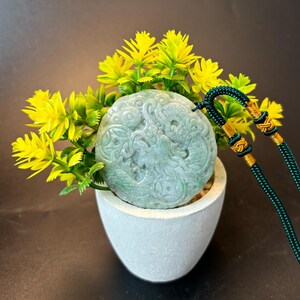May include: A round jade pendant with a carved dragon design. The pendant is green and white and is hanging from a green cord with gold accents.