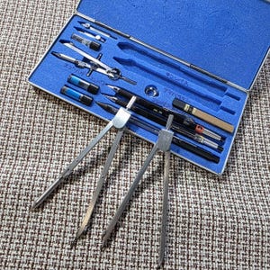 May include: A blue case containing a set of drafting tools, including compasses, dividers, and pens. The tools are silver and black, with some blue accents. The case is open, revealing the tools neatly arranged inside.