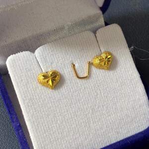 May include: A pair of gold heart-shaped earrings with a textured surface. The earrings are displayed in a blue velvet jewelry box.