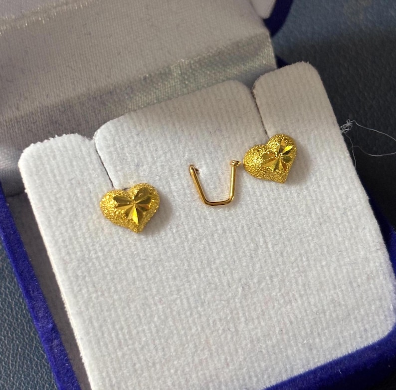 May include: A pair of gold heart-shaped earrings with a textured surface. The earrings are displayed in a blue velvet jewelry box.
