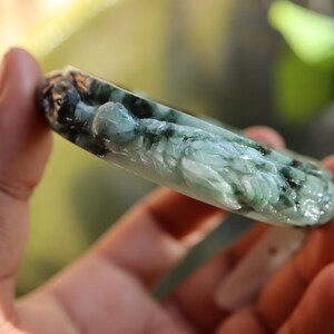 May include: A carved jade bracelet in shades of green and black. The bracelet features a detailed relief carving of a figure. The jade is polished and has a translucent quality, with variations in colour and texture.