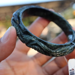 May include: Dark green jade bangle with carved floral or leaf design. The bangle is a circular shape and appears to be made of a solid piece of jade. The carving has a textured appearance. The bangle is held in a hand.